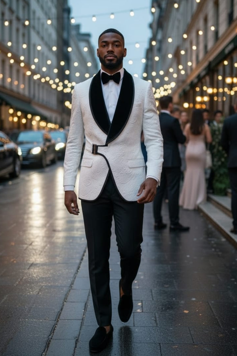 A man walking down a city street at dusk wearing a white tuxedo jacket with black satin lapels, a black bow tie, and black trousers.